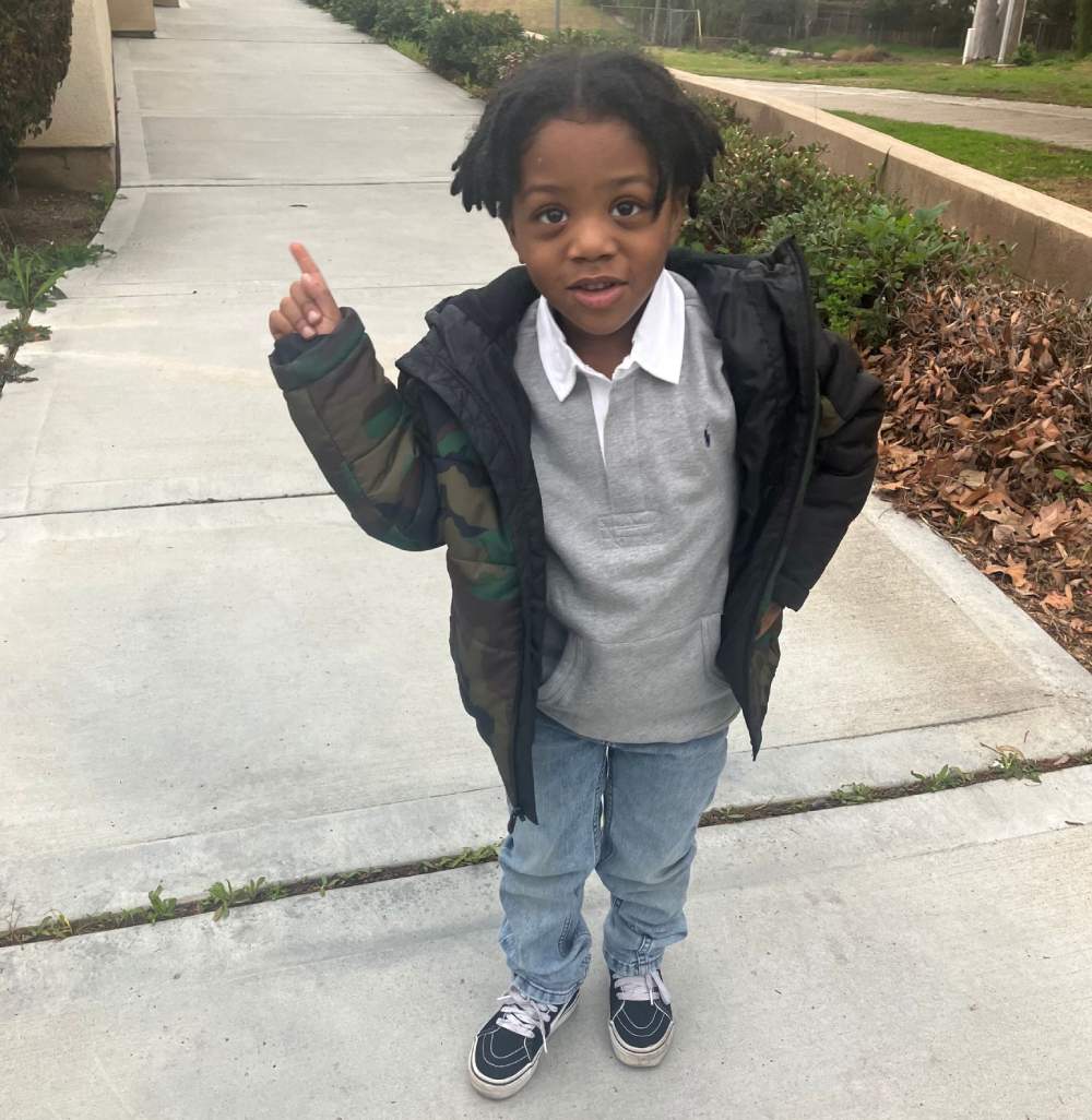 Pictured: A child is wearing blue pants, a grey shirt and a green camouflage jacket. They are holding their right hand up pointing one finger and have their left hand on their hip. The background shows a sidewalk and green bushes.