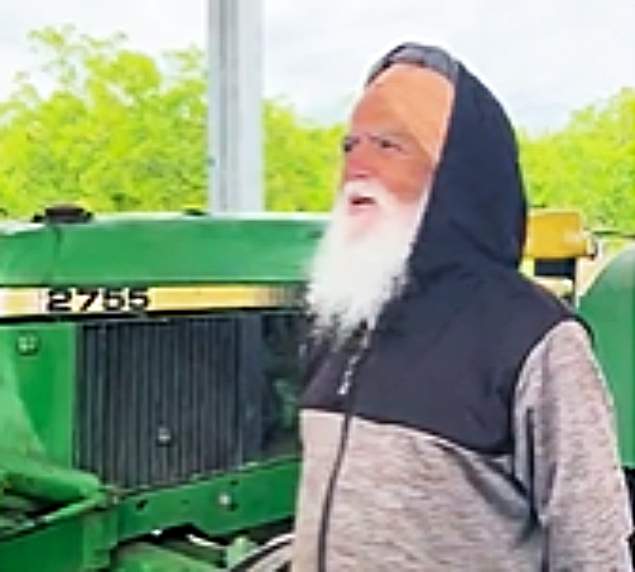 Pictured: A man named Dilbagh Singh is pictured wearing a grey and black jacket in front of a tractor; he has a white beard. The sky is blue and there are trees in the background.