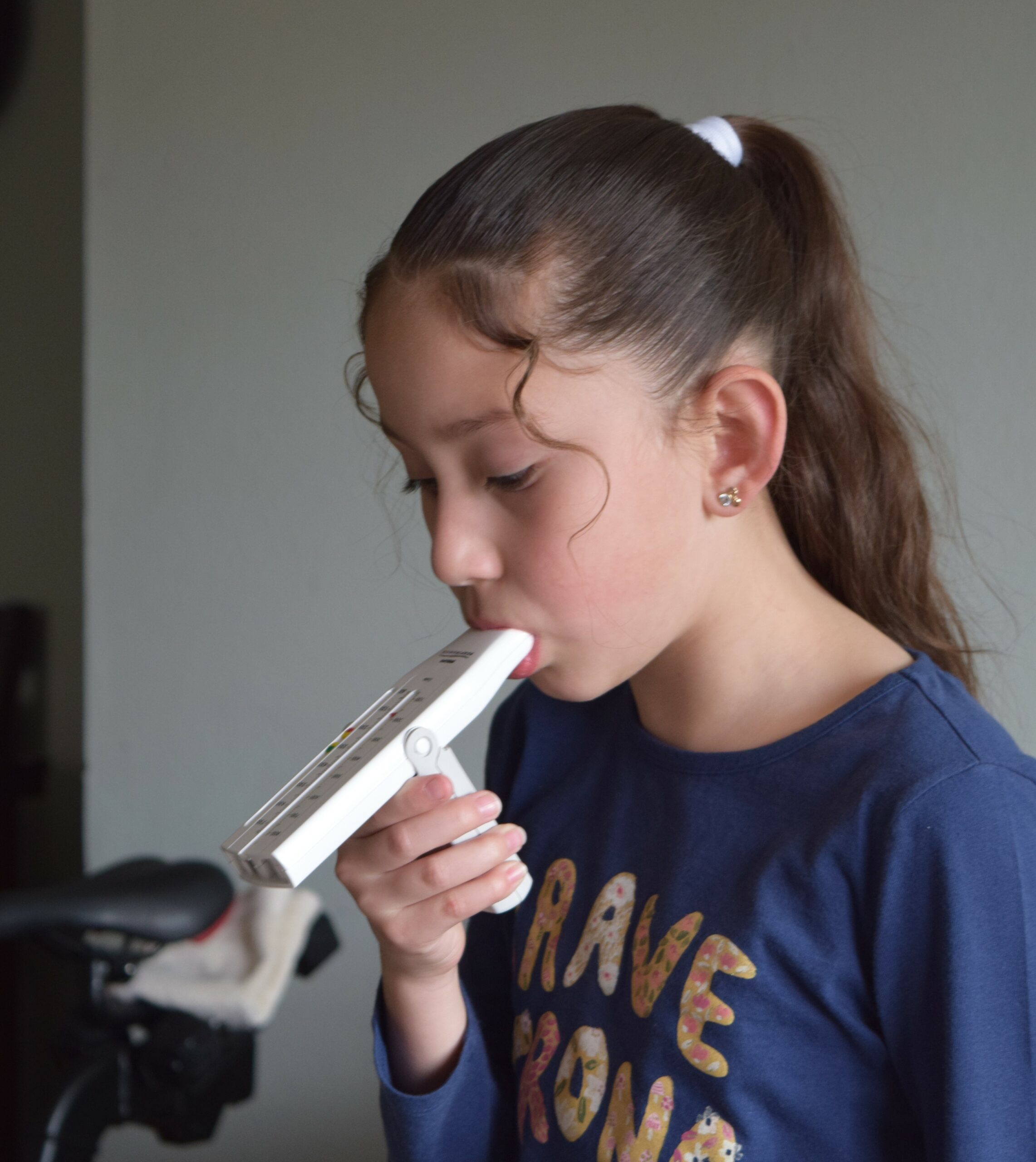 Pictured: A young girl named Melanie is pictured in a blue shirt and using a white peak flow meter. The background shows a bicycle seat and a grey wall.