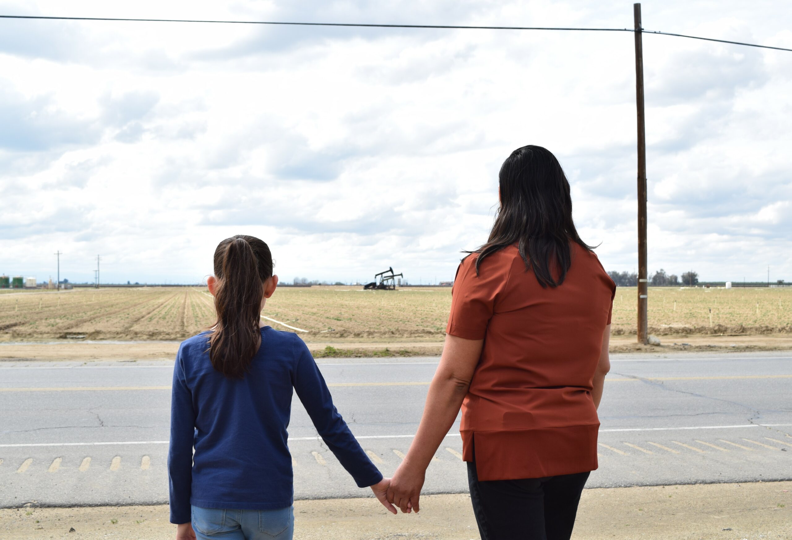 Pictured: A young child is on the left wearing blue with their mother on the right wearing red. They are holding hands and looking out into a large empty field located in Arvin. The sky is cloudy.