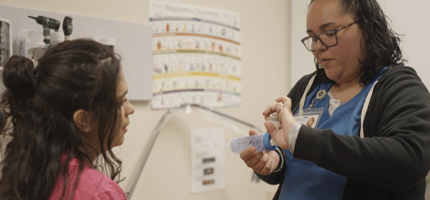 Pictured: A patient wearing a pink shirt is sitting on the left in a medical office. A community health worker stands on the right wearing blue scrubs. The community health worker is demonstrating how to use an asthma spacer. There is a whiteboard and two white posters in the background.