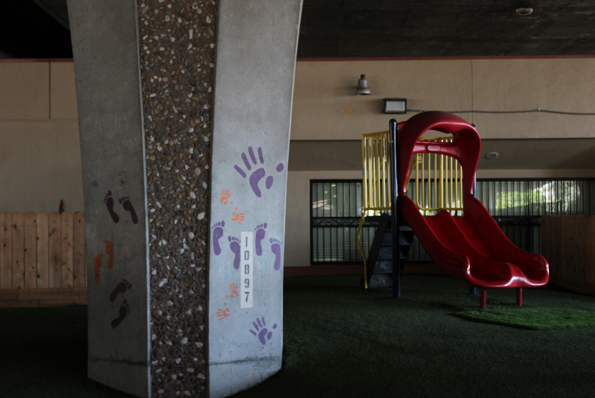 Pictured: A red playground sits behind a grey column that holds up a freeway in South Stockton. The ground is green, and the column has purple and orange handprints and footprints on it. There is very little light.