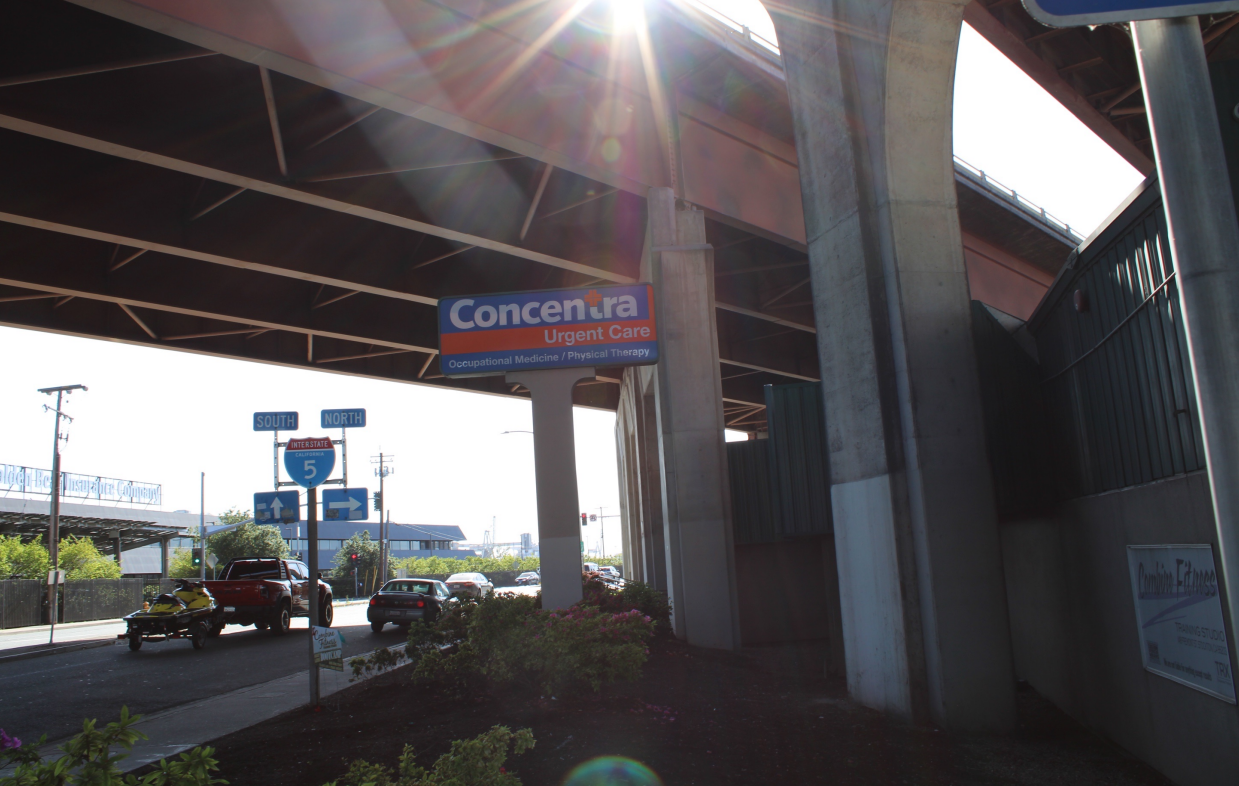 Pictured: A medical clinic is shown underneath a large freeway in South Stockton. There are three cars driving past the clinic. The sky is sunny and bright but beneath the freeway, it is shady and dark.