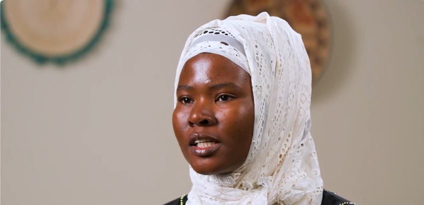 Pictured: A client is shown talking during an interview and wearing an embroidered white hijab. The background is tan and the wall holds two decorative dishes.