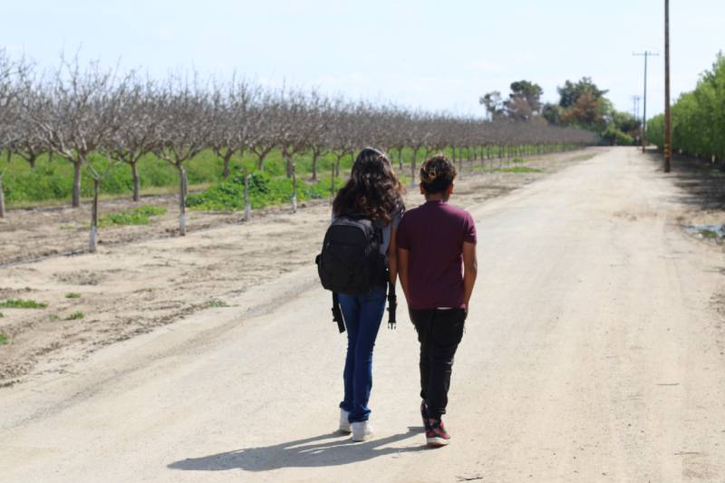 Pictured: Two young students are walking side by side along a dirt path on their way to school in Arvin. On the left, a student wears a backpack and the student on the right wears a dark purple shirt.