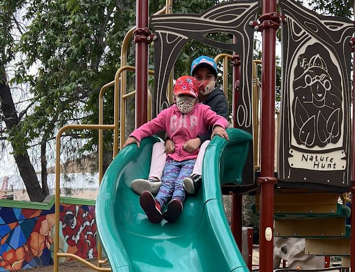 Pictured: Two children are sitting on a green slide together wearing hats and masks. The background shows the rest of the playground structure and large green trees.