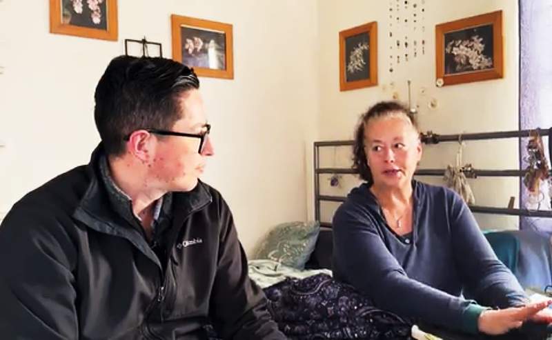 Pictured: A client named Amy sits on the right wearing a dark blue long sleeve shirt. An asthma health management worker sits on the left wearing a black zip-up jacket. The two are sitting on a bed talking. There is a bed frame in the background and four pictures on the wall.