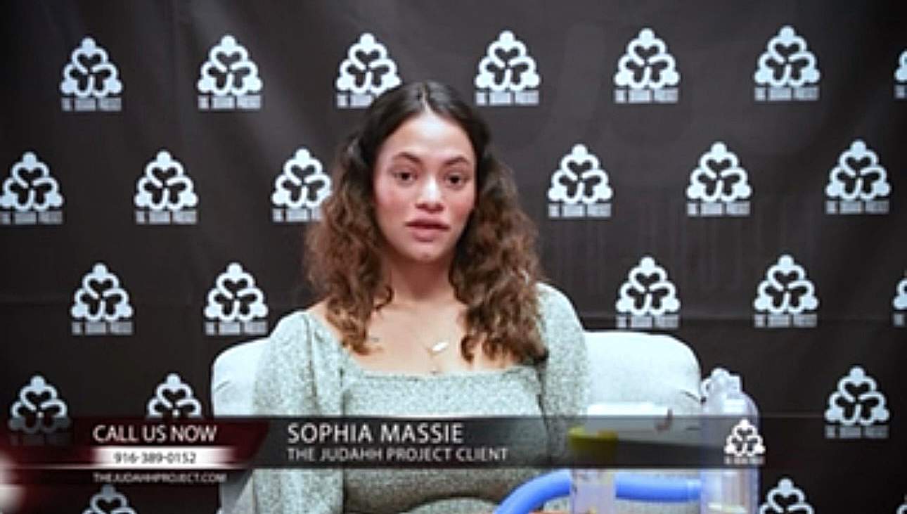Pictured: An adolescent named Sophia Massie is sitting on a chair in front of a black background that has The Judahh Project’s logo. They have curly brown hair and are wearing a light green long-sleeved shirt. They are in the midst of being interviewed.
