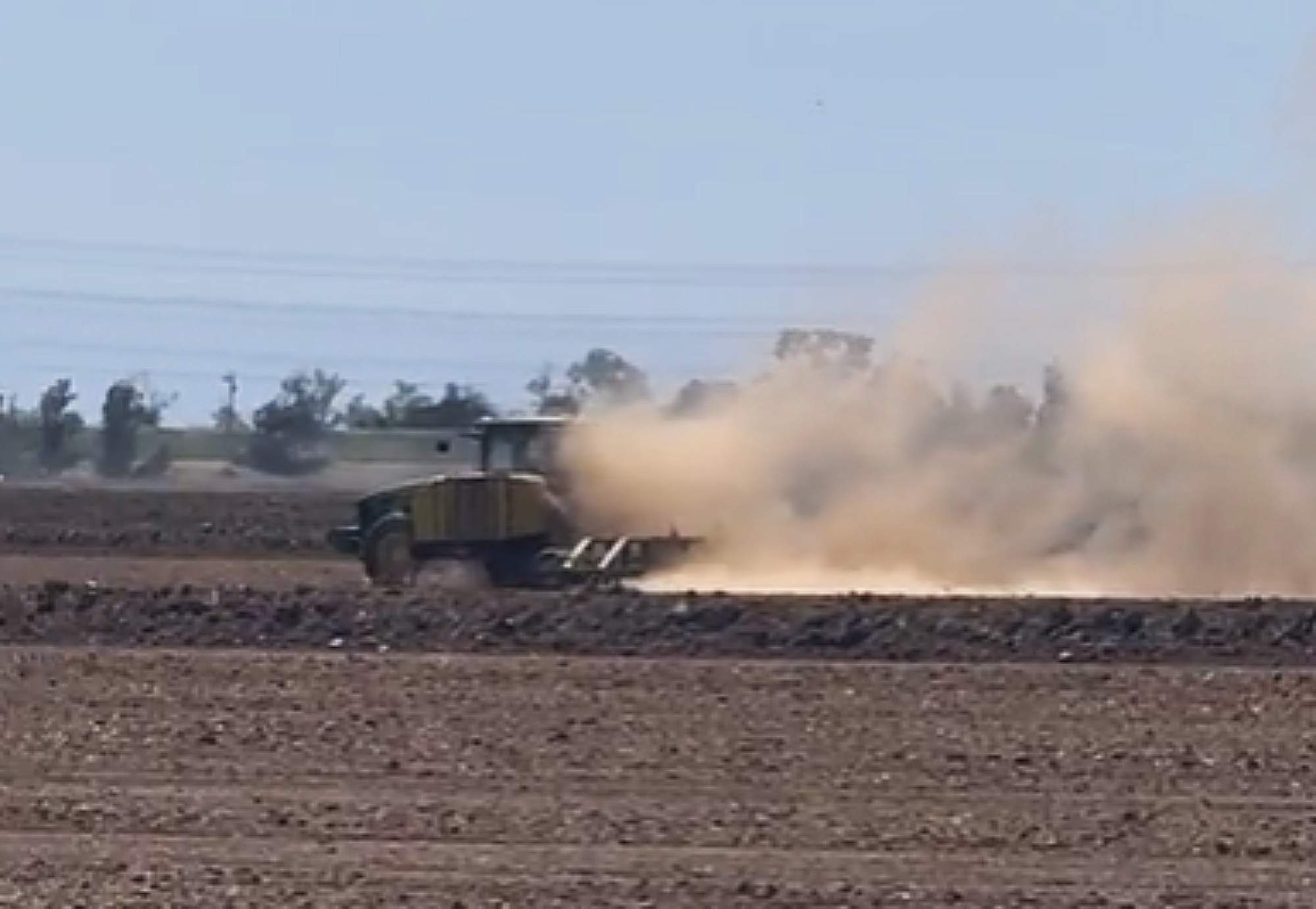 Pictured: A tractor plows through a field of dirt in the daytime of Sutter County. The sky is blue but the large cloud of dust that follows the tractor pollutes the air.